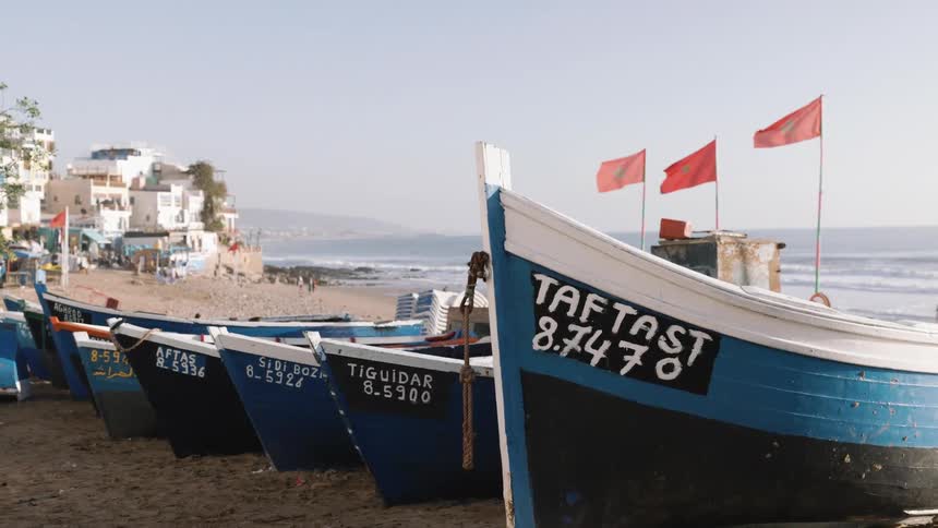Fishing boats lined up on the sand with red flags in the background and the sea on the horizon