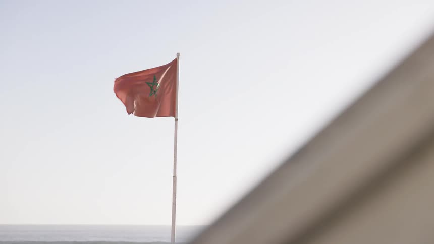 A red moroccan flag bearing a green pentagram flutters atop a slender pole against a pale sunlit sky with the distant ocean faintly visible on the horizon and a blurred architectural edge in the foreground