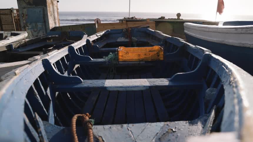 Blue wooden fishing boats sit parked on a sandy shore near the ocean while a small red flag flutters in the background under bright sunlight