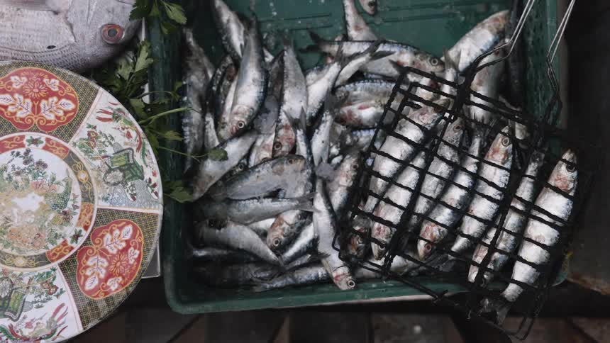 From an overhead view a decorative plate is lifted away from a crate of fresh fish revealing a bowl next to sardines ready for grilling