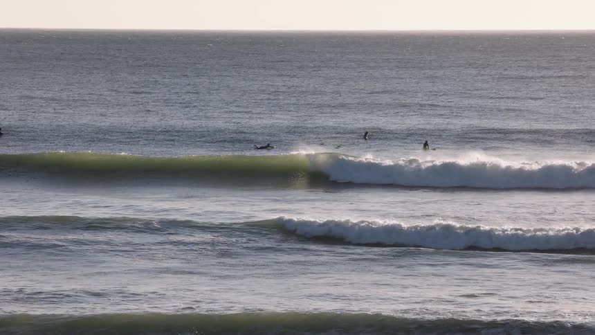Clean evenly spaced ocean swells roll toward shore as a few surfers wait beyond the breaking wave their silhouettes rising and falling against the calm open expanse of the sea