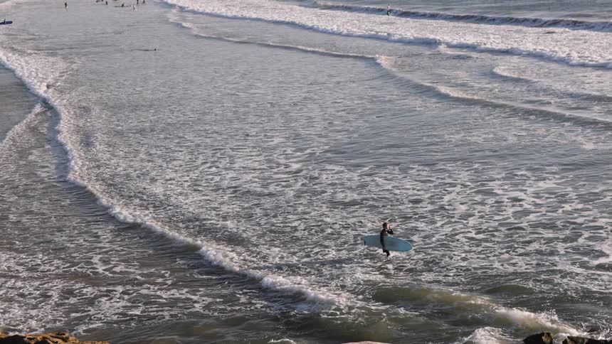 Man on the beach carries his surfboard into the water as waves roll in to shore