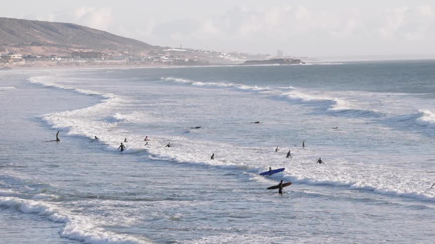 A broad coastal panorama captures numerous surfers scattered across rolling waves with a rugged shoreline and distant hillside town stretching along the horizon under a soft hazy sky
