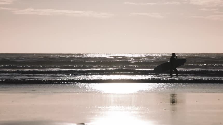 A silhouetted surfer walks across the shallow water of a beach carrying a surfboard under their arm the setting sun reflects off the ocean waves and the wet sand creating a bright and shimmering landscape