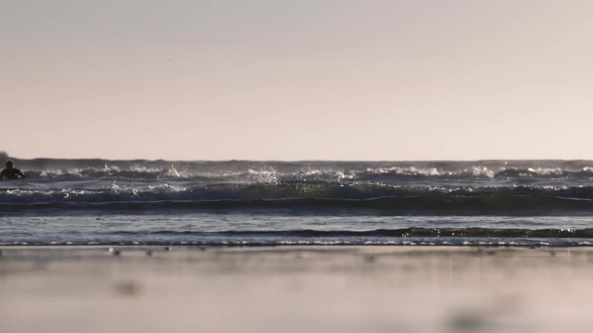 Lowangle shot of ocean waves crashing and rolling onto a shore during a clear sunset or sunrise a silhouette of a person is visible in the distance among the waves as the water flows over the sand