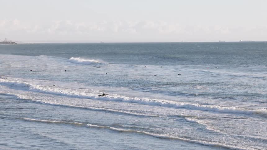 A wide ocean view shows gentle waves rolling toward shore as scattered surfers float beyond the break with one figure standing in the whitewater arms outstretched beneath a pale sky and calm open horizon