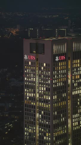 The top of a tall skyscraper with the ubs logo is illuminated against the dark night sky light glows from many office windows as the camera slowly pans out to show the surrounding city lights