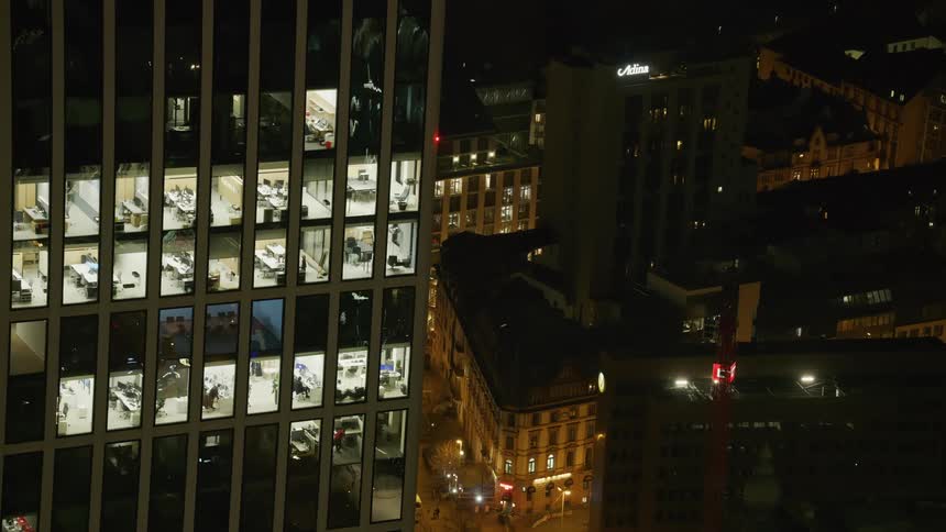 An office building with many lit windows shows people working at their desks late at night the modern skyscraper stands next to older shorter buildings in a brightly lit city