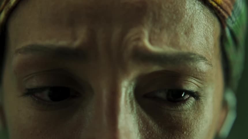 An extreme closeup shows a womans eyes and brow as she looks rapidly side to side with a worried expression and she wears a colorful headscarf