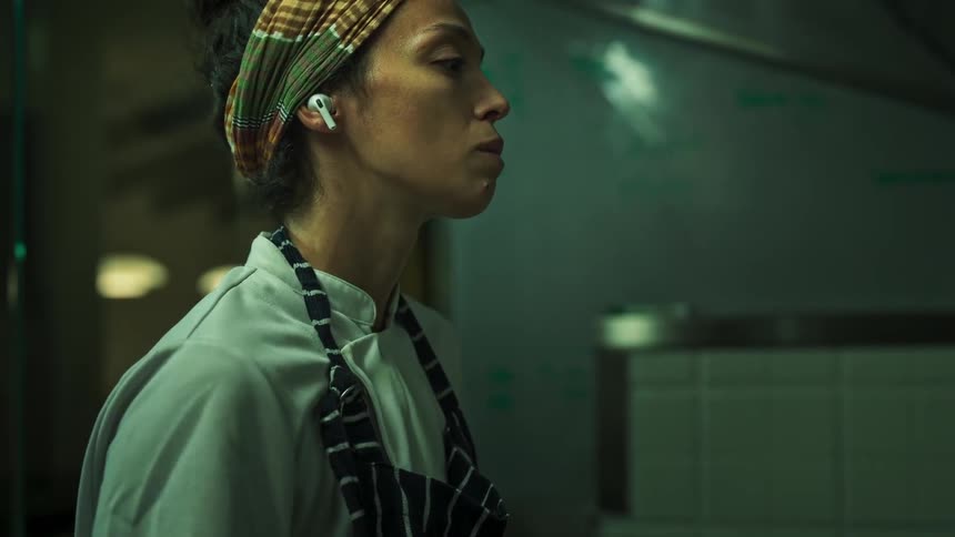 Woman wearing an apron and earbud checks a smartphone in a professional kitchen environment