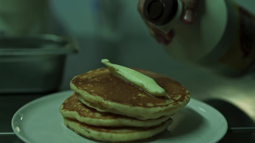 A man uses a knife to spread butter over a stack of three pancakes on a white plate then pours dark maple syrup from a bottle over them before reaching to pick up the dish in a kitchen setting