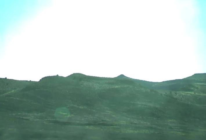 A camera captures a sequence of rolling green hills and mountain ridges under a bright sky filmed through a moving vehicle window which creates occasional light flares and reflections across the natural landscape