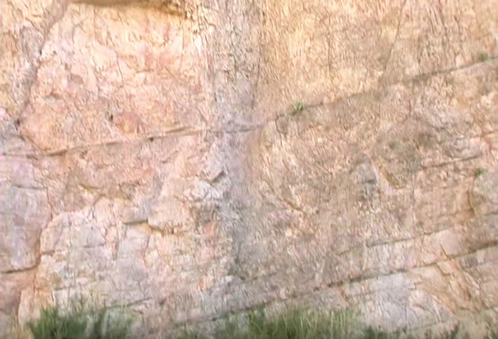 A camera tilts up from a rippling river and grassy bank to reveal a massive layered rock cliff before cutting to a blurry view of a vast desert landscape with mountains in the distance
