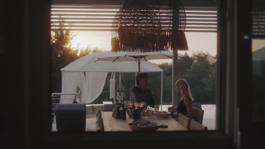 View from inside of a man and woman sitting at a table with candles and food on a patio as he opens a bottle of champagne