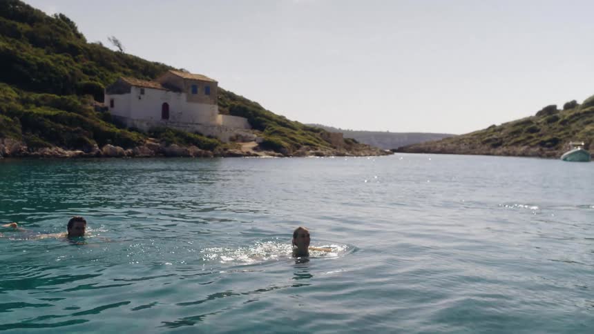 Two people swim in the blue tinted water near a slope holding a large house on a summer day