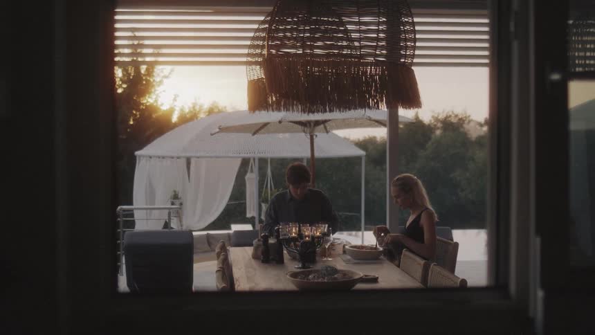 A man and a woman sit at a wooden table on a terrace at sunset eating a meal together by candlelight while a wicker lamp hangs above them and a pool sits in the background