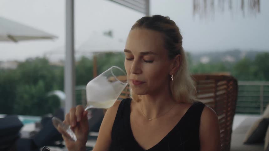 A blonde woman in a black dress sits at an outdoor terrace table lifts a glass of white wine to take a sip tastes it with a thoughtful expression and then continues talking to someone offcamera as evening falls over a pool in the background