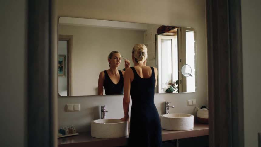 Woman applies makeup in front of a mirror in a residential bathroom with two sinks