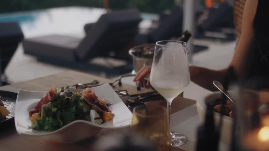 A woman uses metal tongs to serve a guest fresh salad with cured meat onto a modern white plate on an outdoor table beside a glass of white wine and small plated sides in a relaxed upscale dining setting
