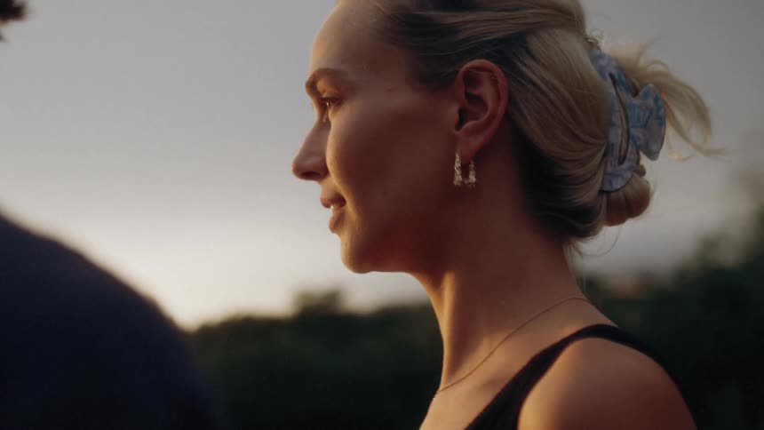 A blonde woman looks lovingly at a man in the foreground her face illuminated by the warm glow of sunset as she smiles and laughs