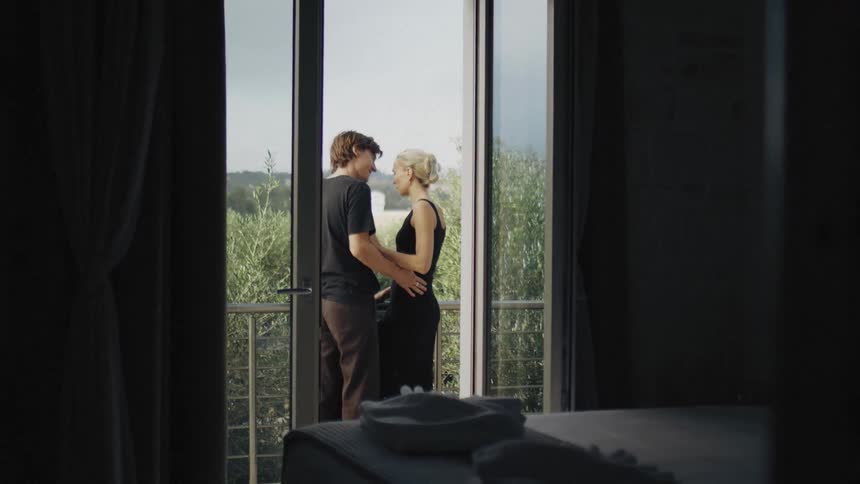 A man and a woman wearing black clothes embrace and kiss on a balcony before looking out at a lush green landscape from a dark room during the day