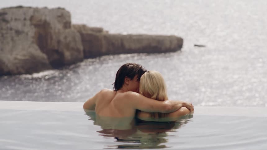 A couple embraces in an infinity pool looking out at the sun dappled ocean and distant cliffs
