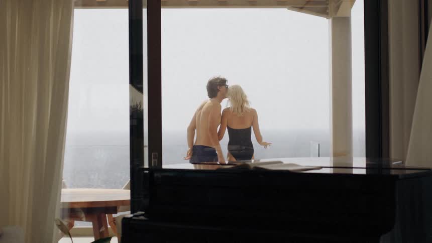 A man holds and kisses a woman while standing on a balcony overlooking the ocean