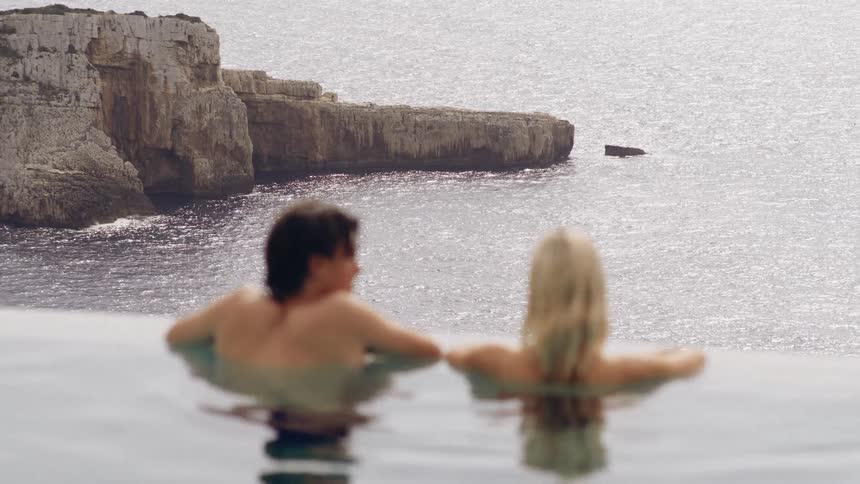 Two people rest side by side in an infinity pool quietly facing the open sea and rocky coastline beyond