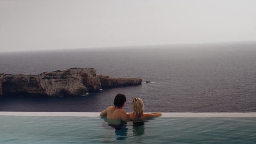 Couple rests against the edge of an infinity pool overlooking the sea and rocky formations