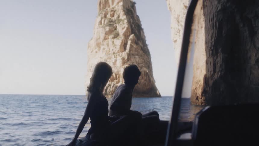 Two people sit happily silhouetted in a small boat inside a sea cave gazing out toward sunlit rock formations rising from calm blue water
