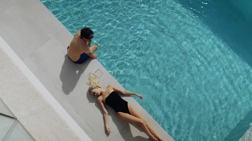 Woman lying near the swimming pool moves her hand through the water while a man sits with his feet in the water on a sunny summer day