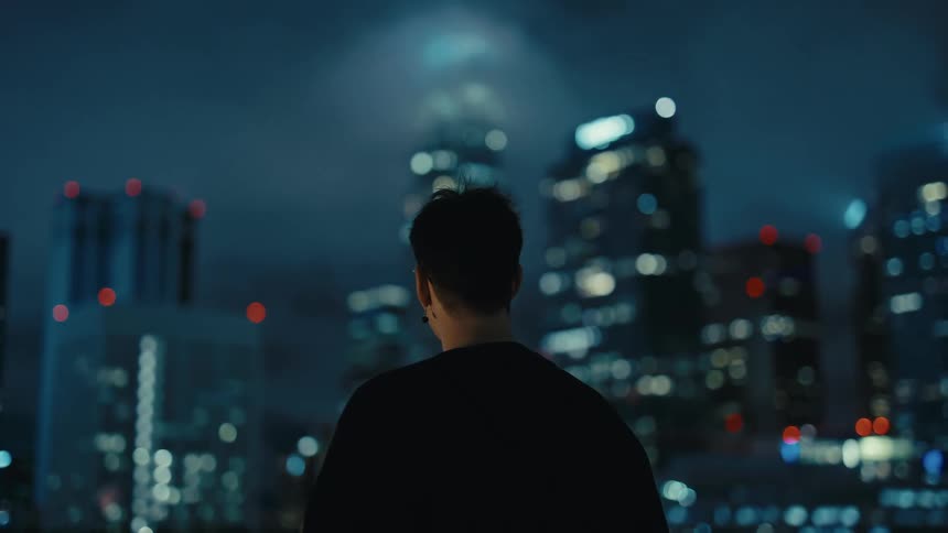 A man with glasses and a black tshirt stands on a rooftop at night looking out at a blurred city skyline filled with glowing office buildings and skyscrapers under a dark sky