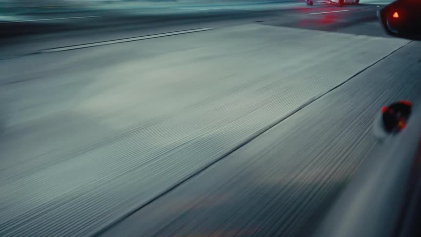A lowangle shot from a moving car captures the textured highway pavement rushing by at night while other vehicles with glowing red tail lights travel ahead under the dark sky