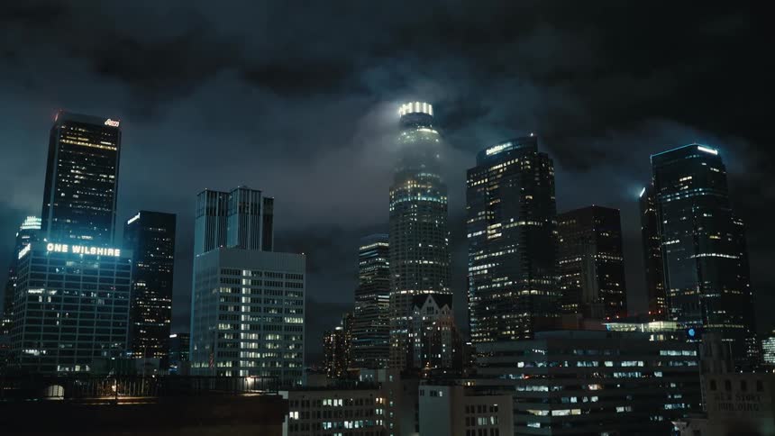Low clouds drift over a brightly lit city skyline at night with tall glass skyscrapers glowing against the dark sky