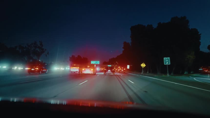 Several cars and motorcycles traveling south on a multilane highway at night passing an exit sign while headlights and street lights illuminate the dark roadway