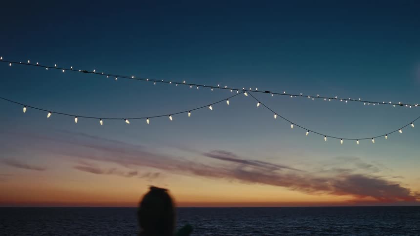 A person walks under white lights strung along an ocean coast at dusk