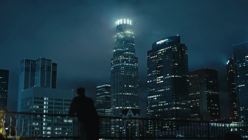 A young man walks across a rooftop balcony in a metropolis at night