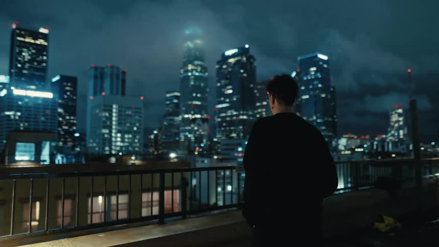 Man walking on a rooftop looking at downtown buildings with lights glowing in the darkness under a cloudy sky