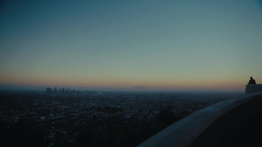 At dusk a lone person stands on a buildings edge gazing over the vast city as it fades into the horizon beneath a quiet sky