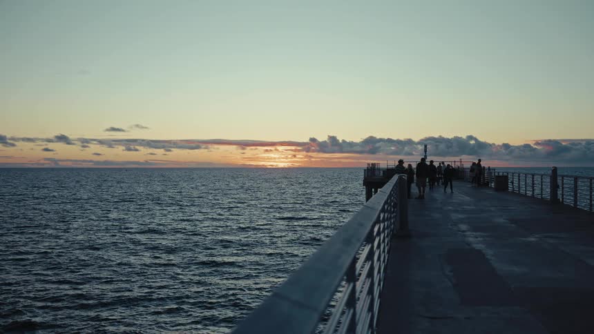 People walk along a concrete pier at dusk as the sun sets over the ocean horizon casting a golden glow through scattered clouds while dark waves ripple below the metal railings of the walkway