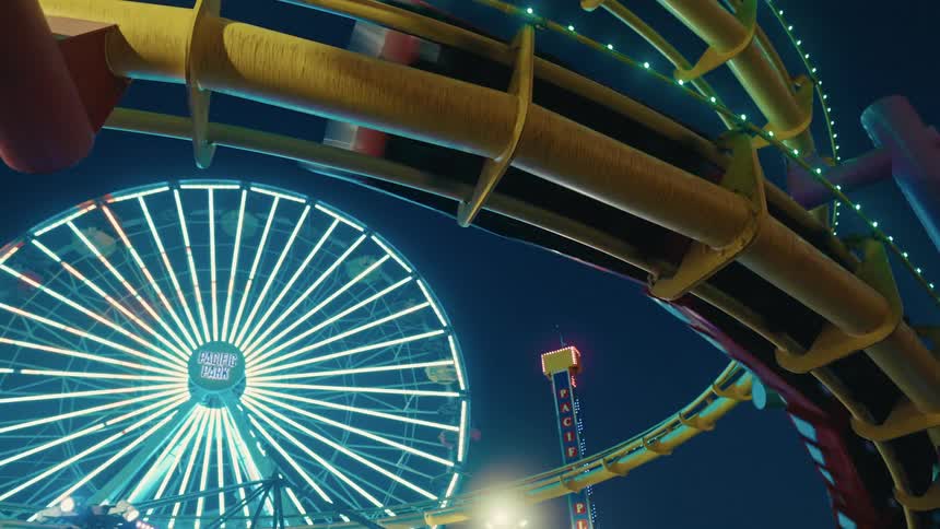 Roller coaster structure in the foreground with a lit ferris wheel at a nighttime amusement park