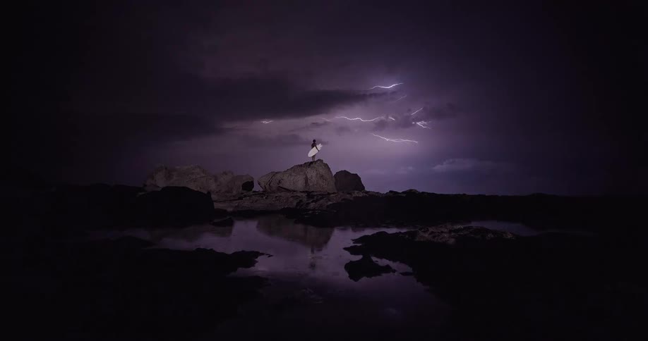 A person holds a white surfboard while standing on a rocky coast during a lightning storm