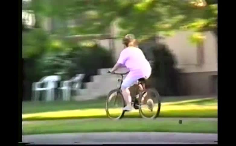 A person humorously rides a bicycle at high speed through a suburban street while lying stretched out on the seat one leg lifted behind them in a playful retro homevideo style shot