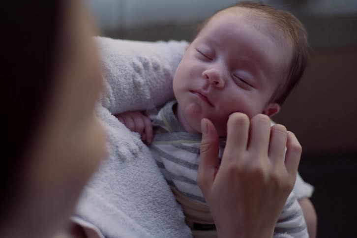 Baby sleeps in an adults arms while a hand gently touches the face