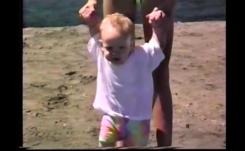 A mother in a striped swimsuit holds a toddlers hands to help him walk across a sandy beach near a blue canopy and folding chairs on a sunny day