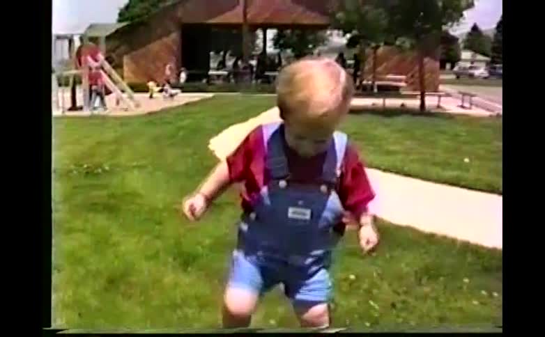 A young blonde boy wearing red shirt and blue denim overalls walks across a bright green lawn in a park with trees toys a basketball court and mountains in the background during a sunny day