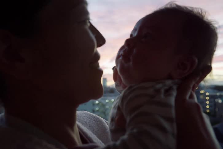 Adult holds a baby face to face near a window with an evening sky in the background
