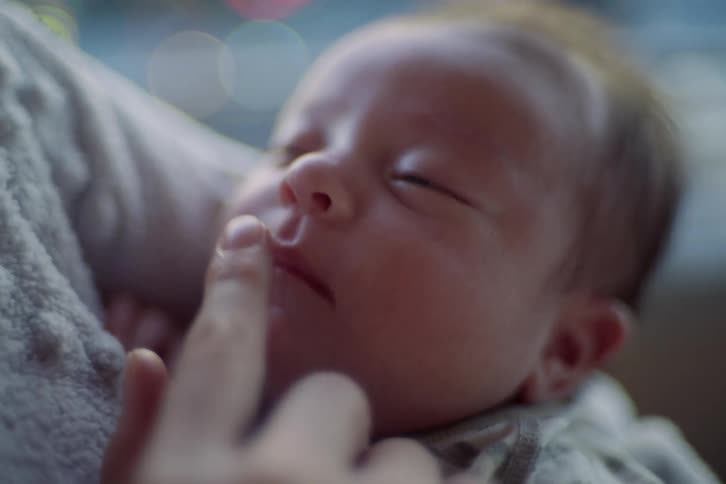 A closeup shows a newborn resting against a caregiver while a gentle finger softly touches the babys nose the infants halfopen eyes calm in warm blurred light