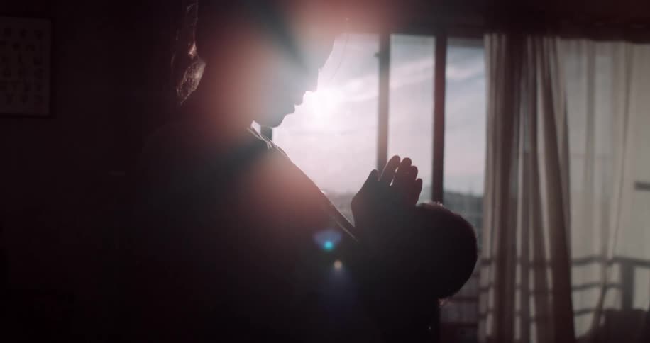 In soft backlight by a window a silhouetted adult shields the sun with one hand while gently cradling a newborn creating a quiet intimate moment
