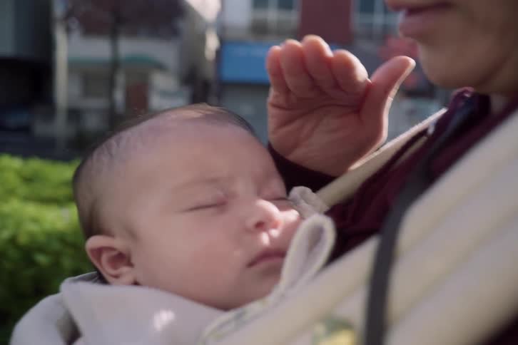 A baby sleeps peacefully in a front carrier outdoors while a caregivers hand gently shields the infants face from the sunlight